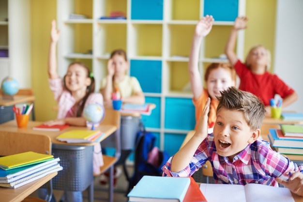 Children in classroom with theirs hands up